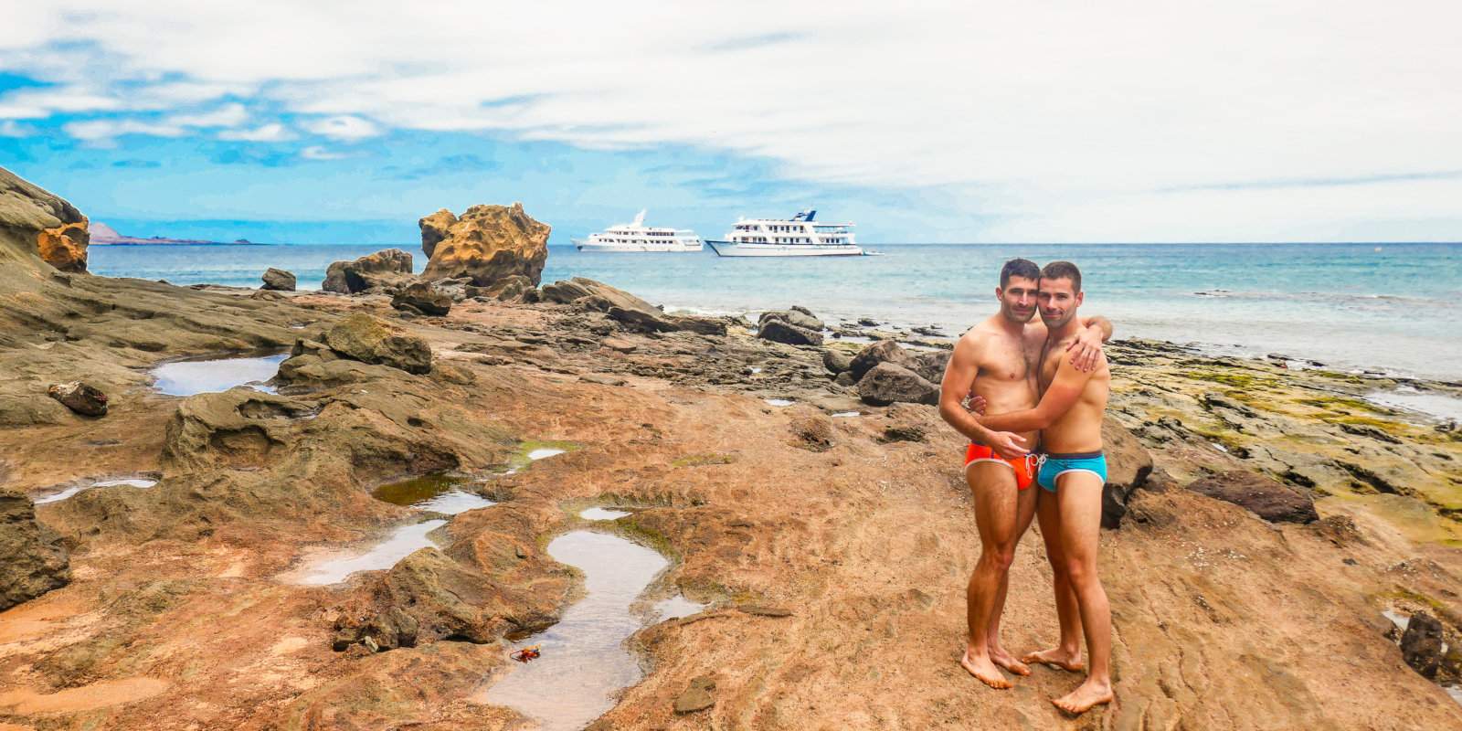 Two men embracing in swimwear on rocks with cruise ships in the background.