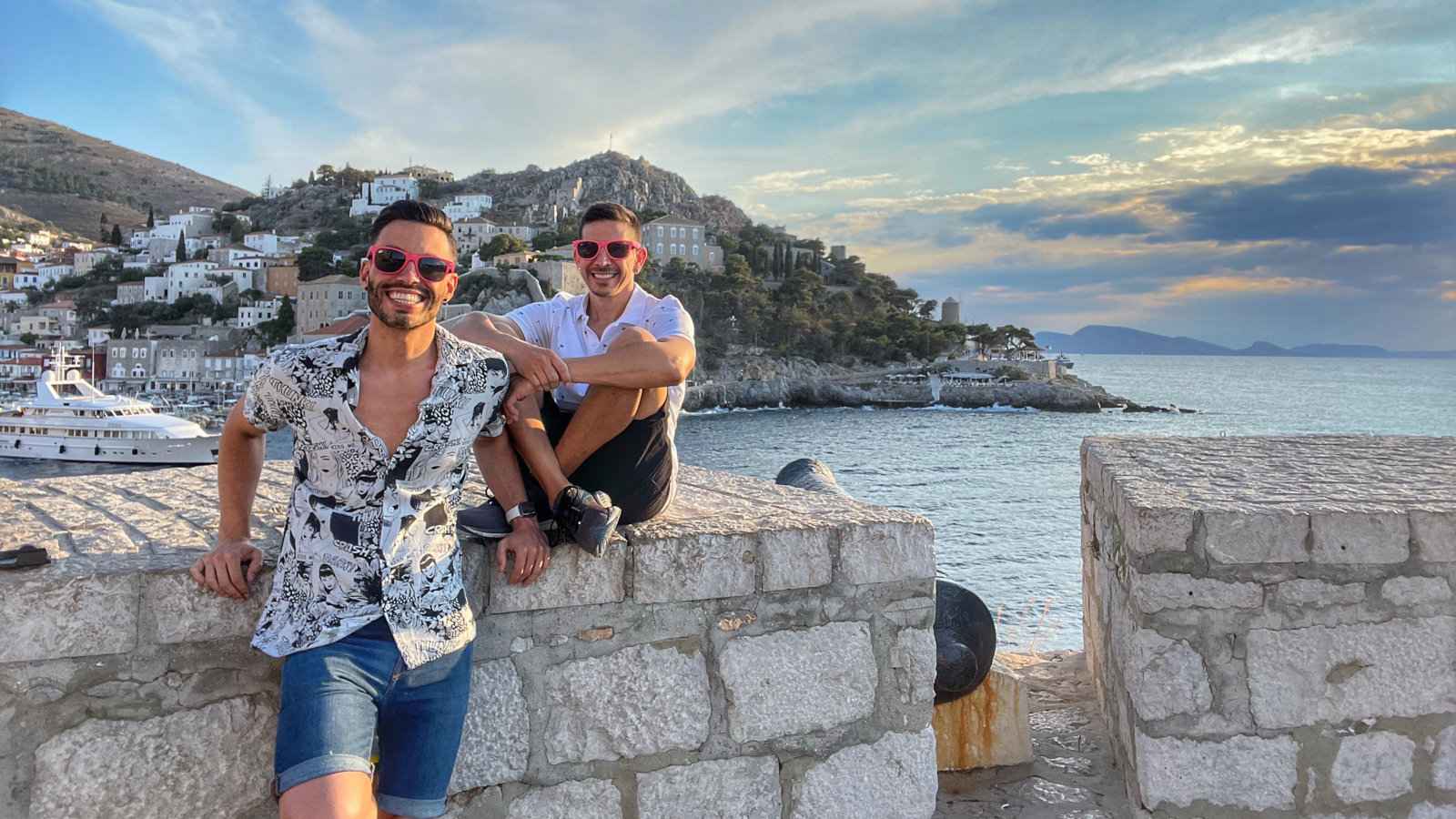 Two men in shorts, shirts, pink sunnies and cheesy grins posing on a stone wall in front of a beautiful bay and harbour town.