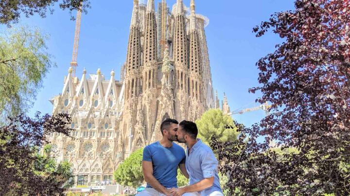A gay couple sitting in front of the Sagrada Familia in Barcelona and sharing a kiss.