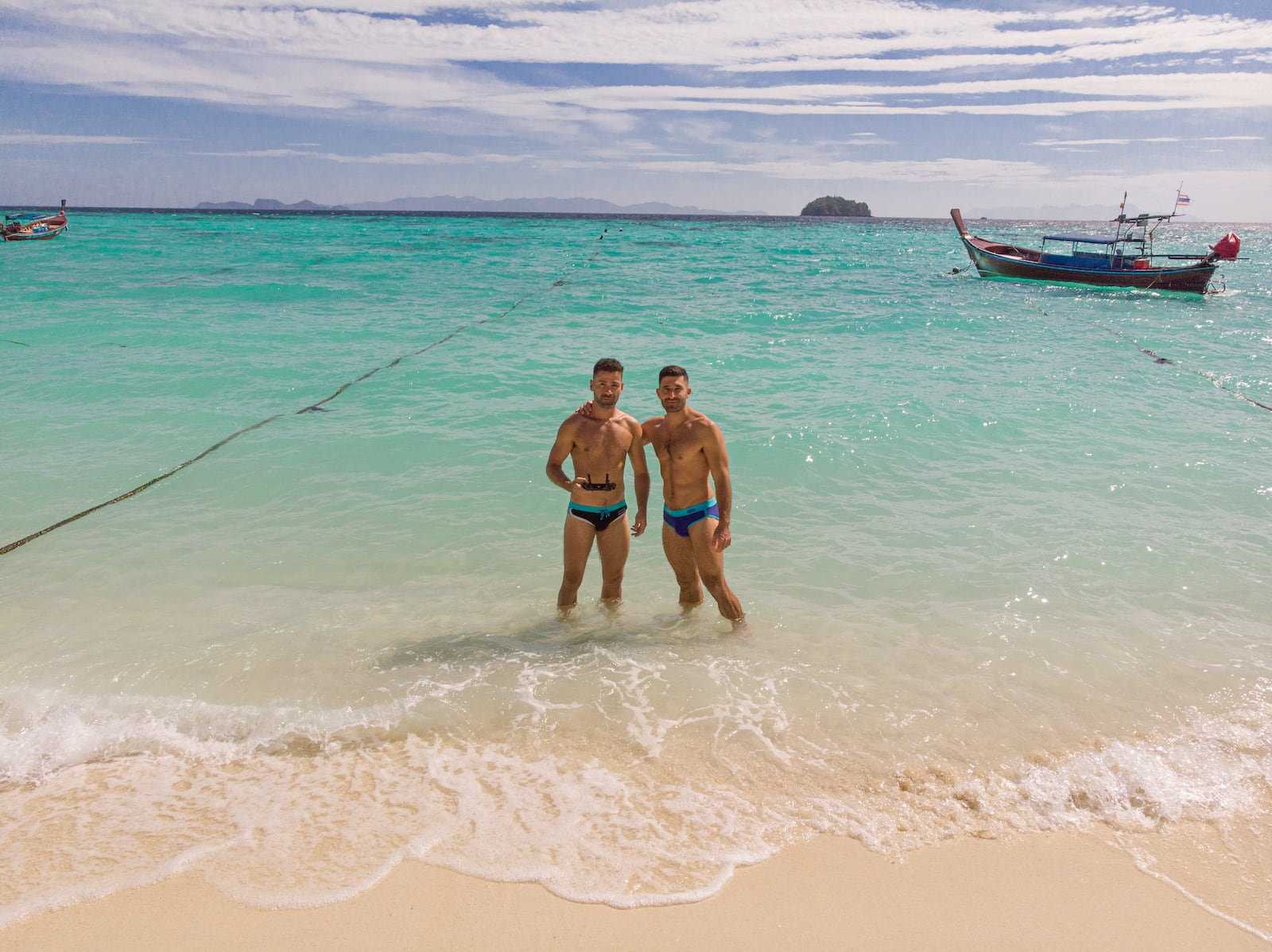 A gay couple in shorts standing in shallow waters with a boat behind them.