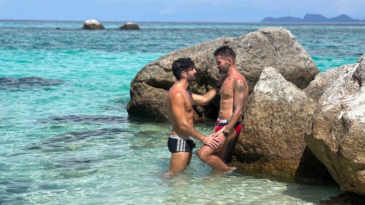 A gay couple in shorts standing in crystal clear waters by some rocks.