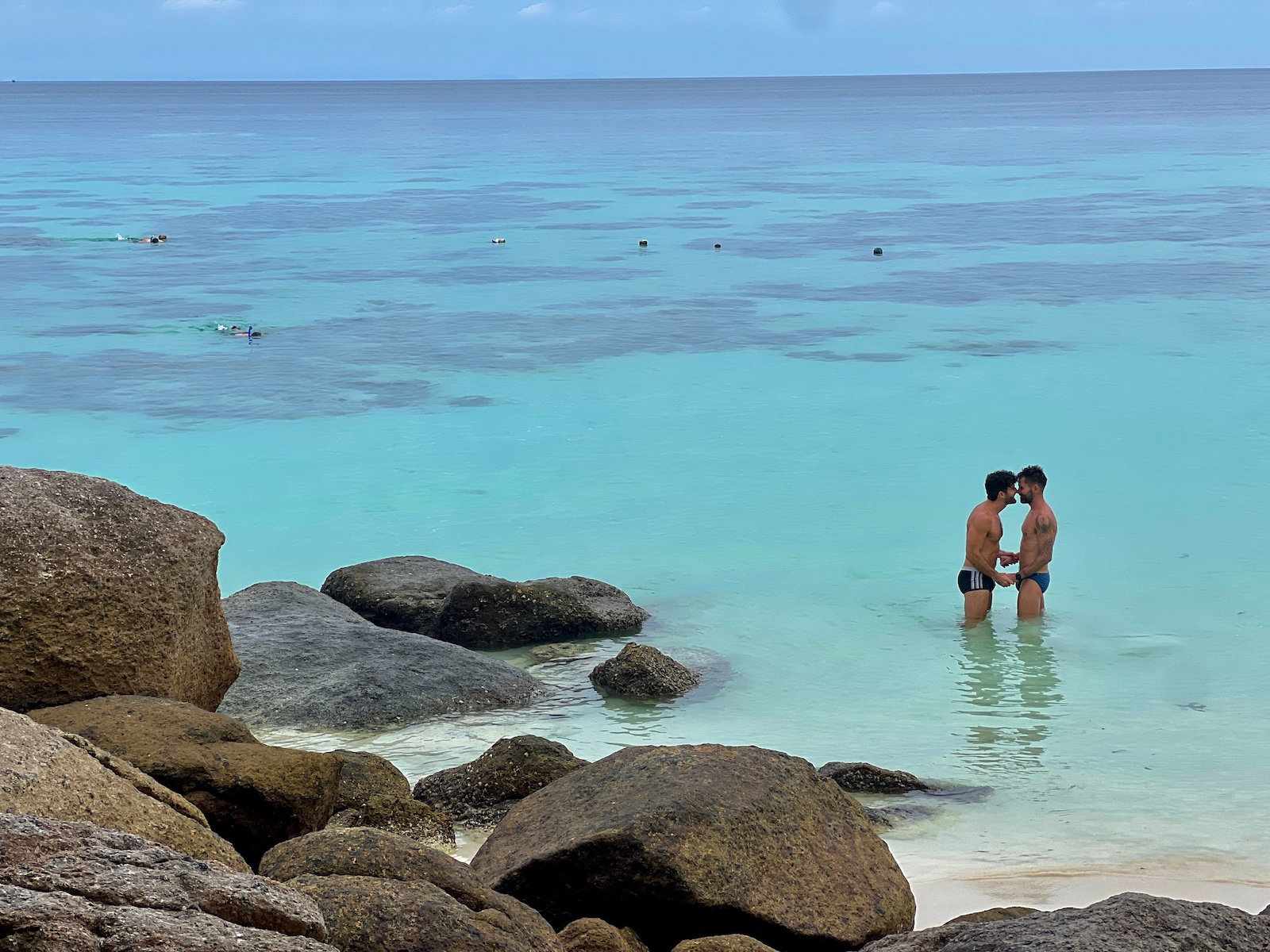 A gay couple standing in azure waters with rocks in the foreground and not another soul in sight.