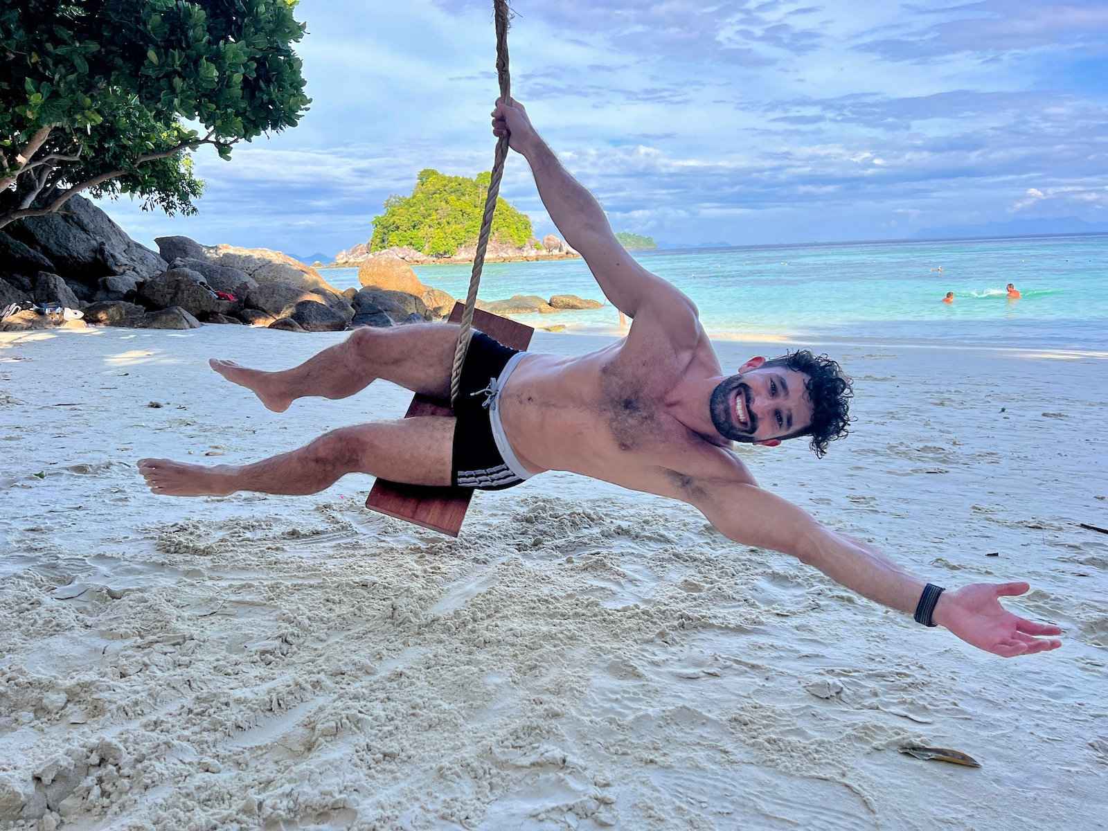 A man in tiny shorts smiling and swinging on a simple beach swing.