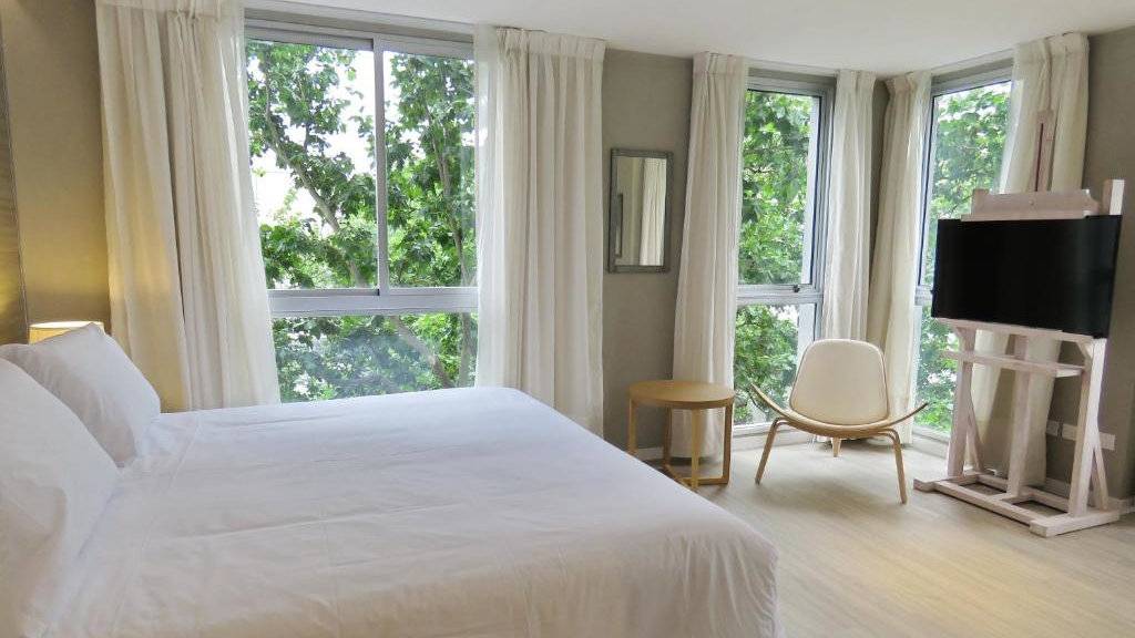 A spacious and light bedroom at Smart Hotel Montevideo with green tree leaves out all three windows.