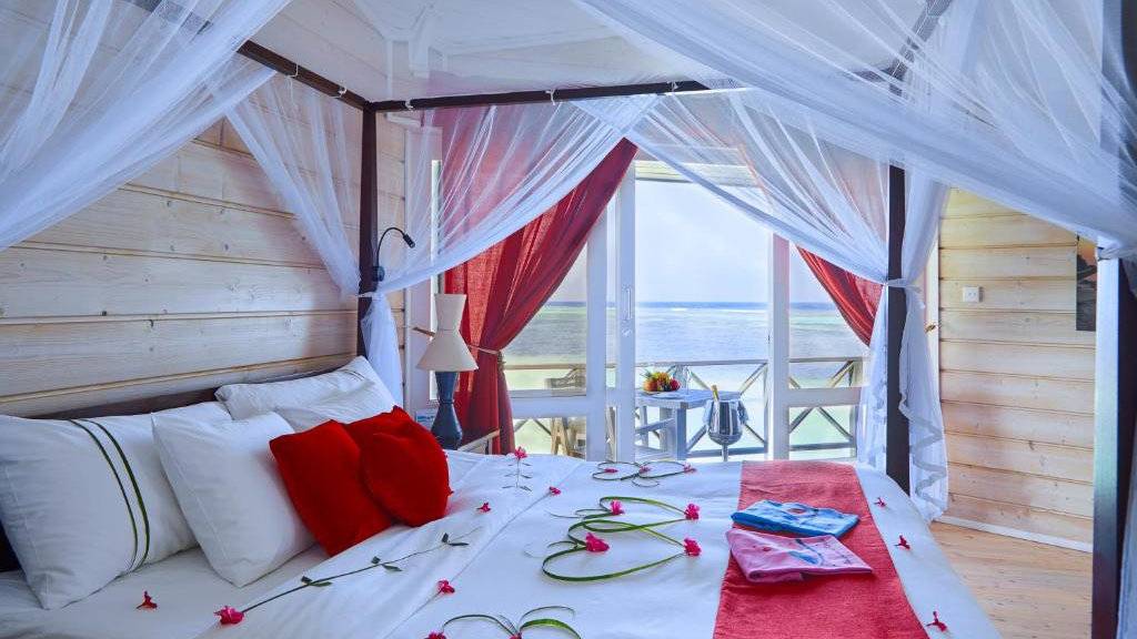 An airy bedroom with flower petals and hearts on a bed with a view from the balcony to the beach in the Maldives.