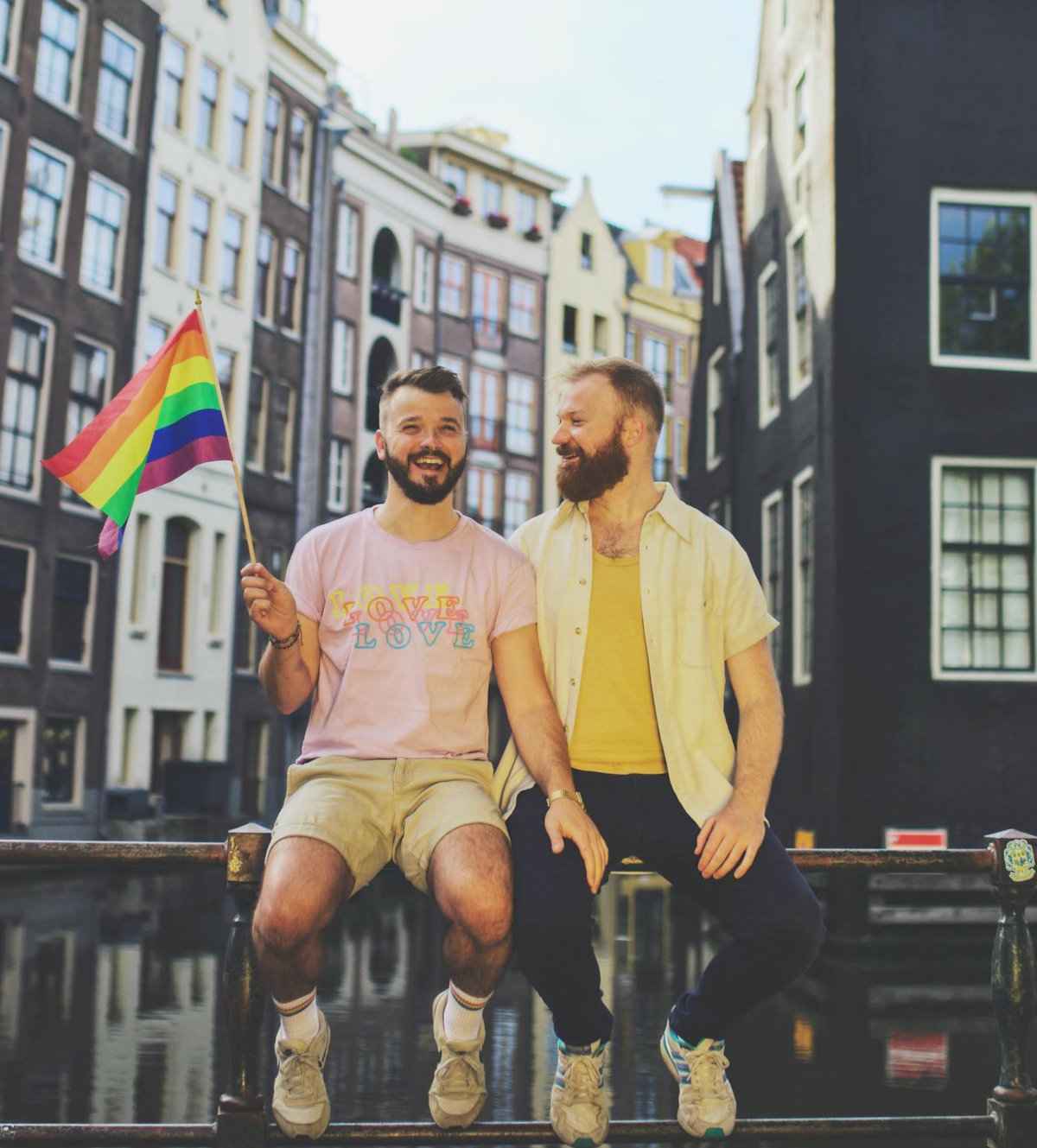 Karl and Daan of Couple of Men sitting next to a canal in Amsterdam while waving a rainbow flag.
