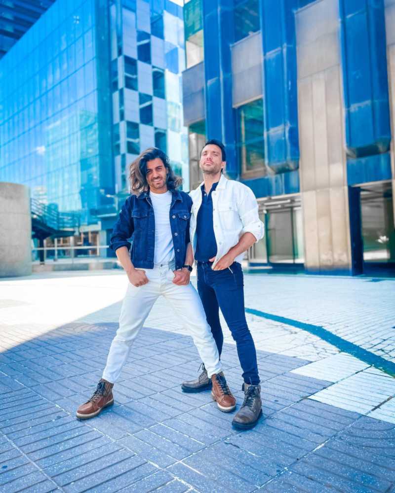 Two men in blue and white denim posing in front of a blue cityscape.