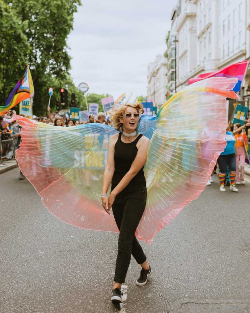 A blonde girl wearing black clothing and a rainbow cape that looks like wings walking down a road.