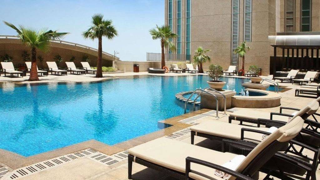 An outdoor swimming pool on a sunny day surrounded by loungers and a tall building.