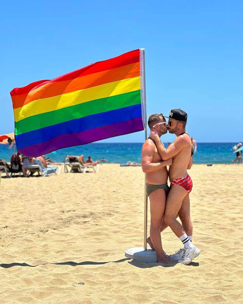 Two gay guys in speedos sharing a kiss on a beach next to a rainbow pride flag.