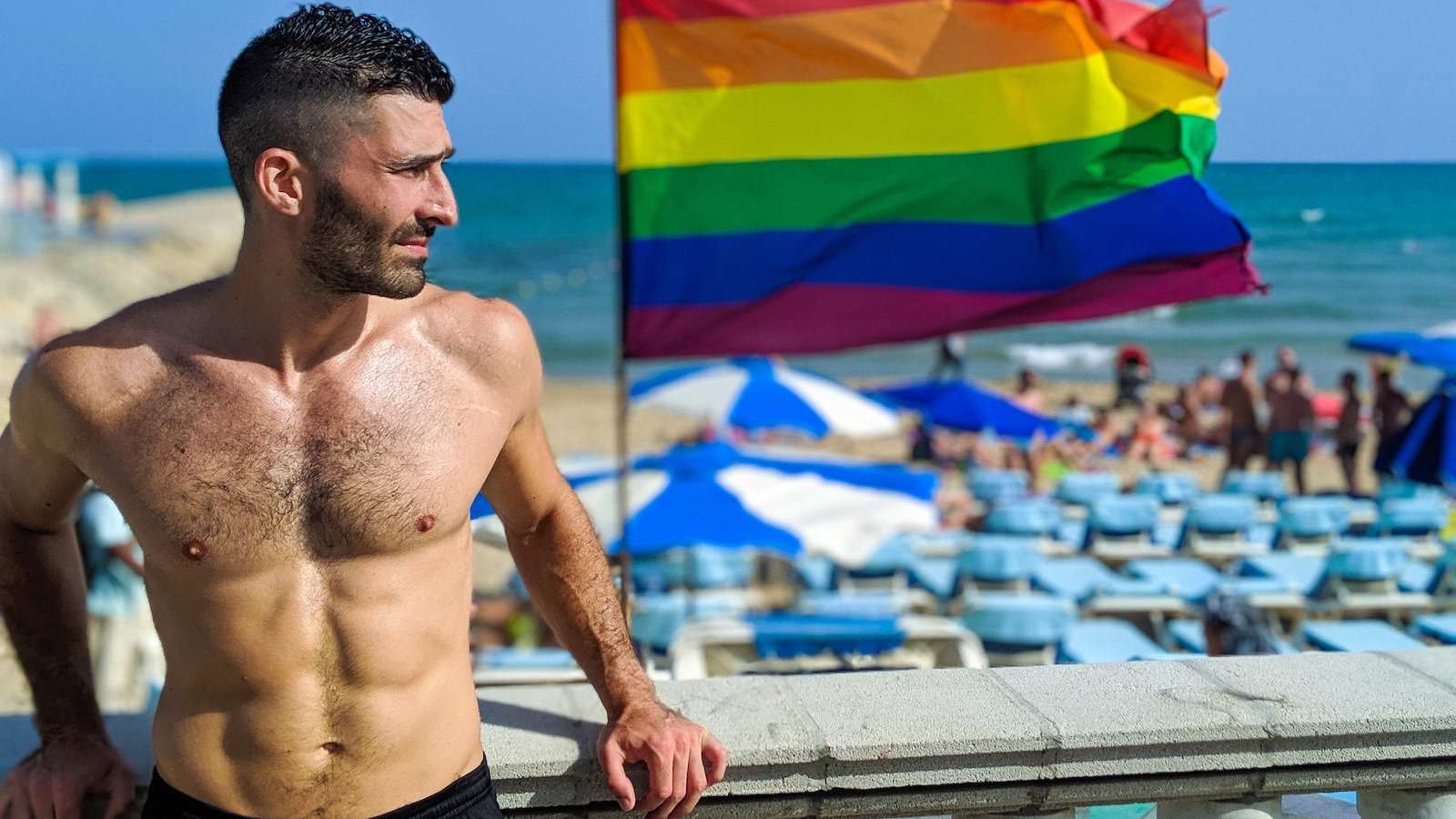 Stefan from Nomadic Boys in front of the gay beach in Maspalomas, Gran Canaria with the gay flag in the background.