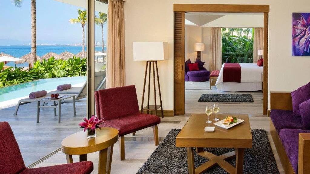 A bright villa sitting room looking into a bedroom with a balcony and private pool outside.