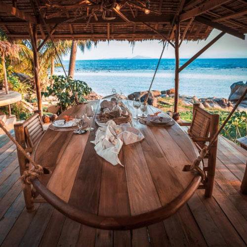 A wooden dining table under thatched roof directly next to a beautiful secluded beach.