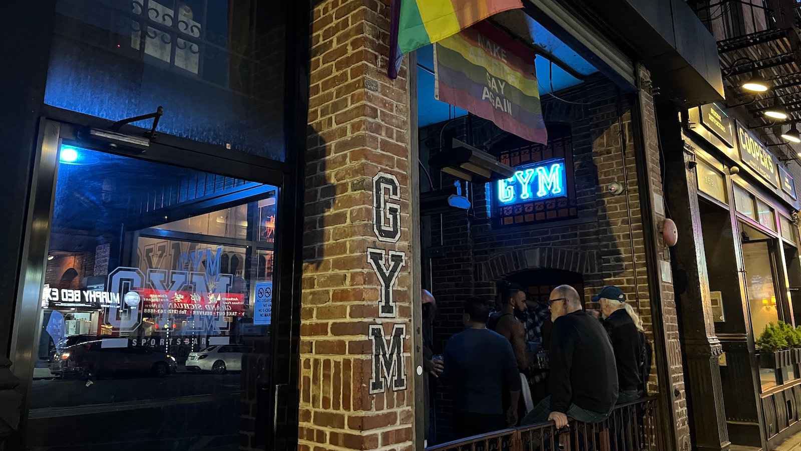 Some patrons sitting at the entrance to Gym gay bar in New York City, with rainbow flags flying.