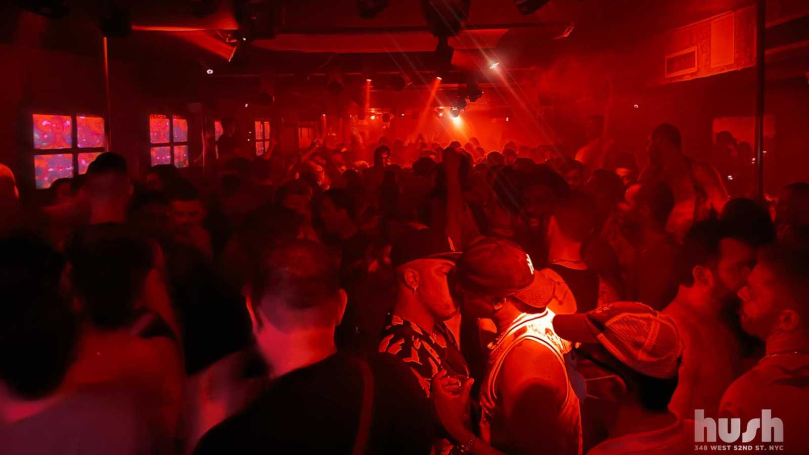 A crowded dance floor lit by red lights showing gay men dancing and kissing at HUSH gay bar in New York City.