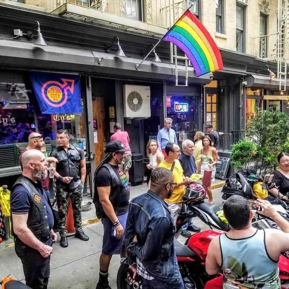 A group of men standing around motorbikes outside Ty's Bar in New York City, with rainbow flags proudly waving.