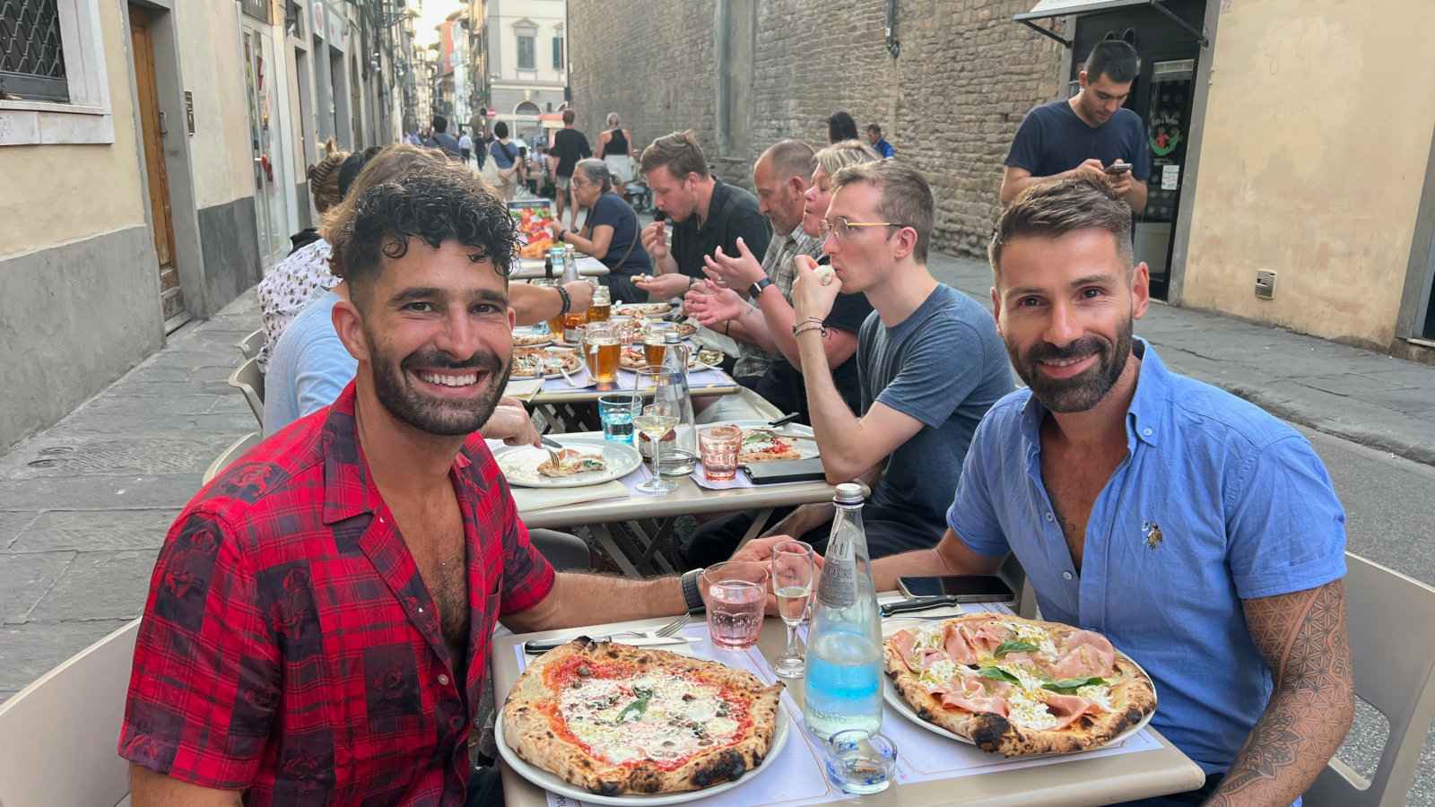Stefan and Seby sitting and smiling at outdoor tables in Florence with pizzas in front of them.