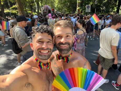 Stefan and Seby posing in rainbow bow ties and a rainbow fan at a park with more people and rainbow flags behind them.