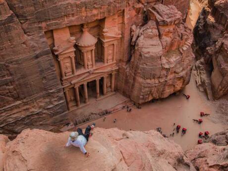 An overhead shot of a man sitting on a rock ledge looking down into a canyon with a building carved into the red rocks.