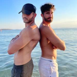 Two shirtless guys standing back to back in front of crystal clear waters.