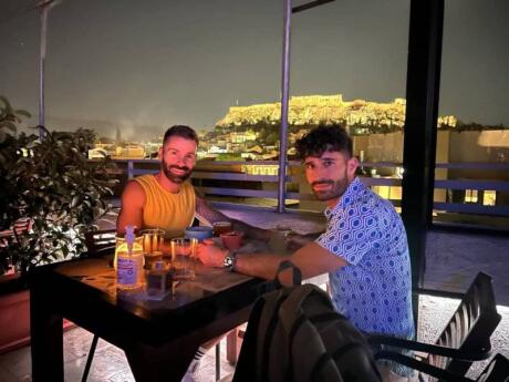Stefan and Seby of Nomadic Boys posing for a photo at a rooftop restaurant at night with the Acropolis lit up behind them.