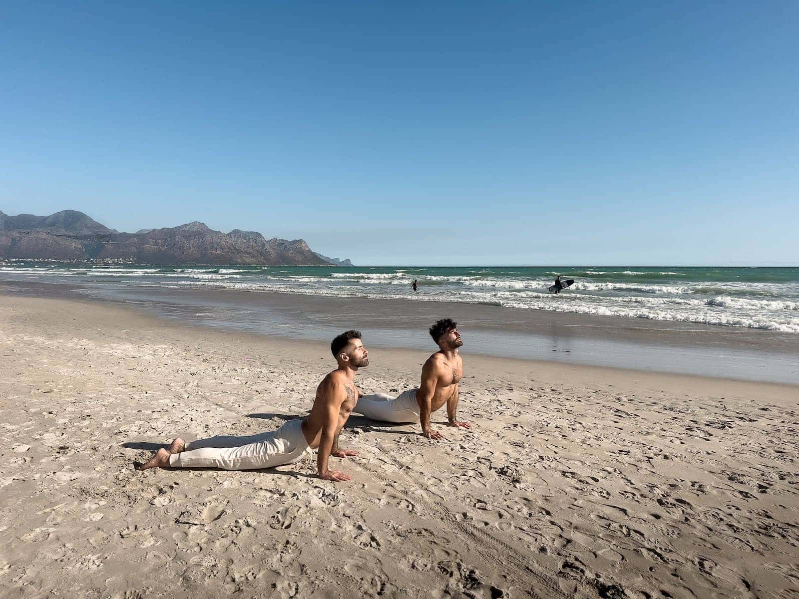 Gay couple doing an upward dog yoga pose on the beach.