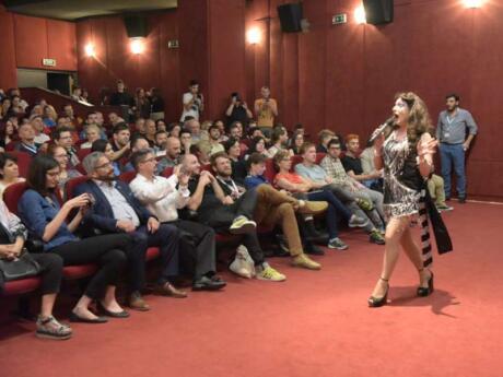 A drag queen talking into a microphone to a crowded movie theater at Outview Film Festival in Athens.