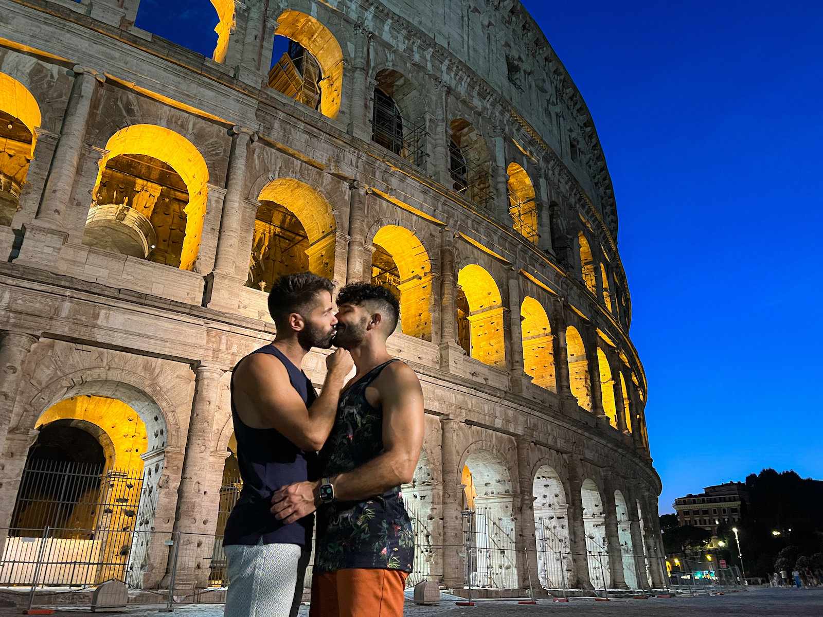 The Nomadic Boys kissing in front of the Colosseum in Rome at dusk, with the arches of the Colosseum lit up.