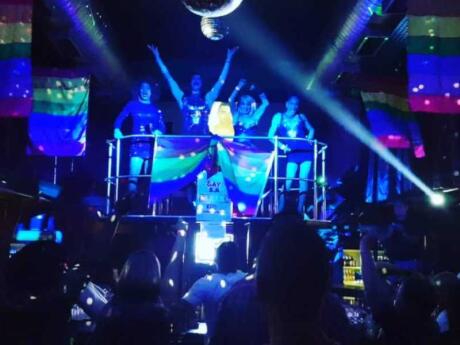 A dance floor lit up with blue and rainbow flags with dancers on a stage above the main crowd at the gay club SA.