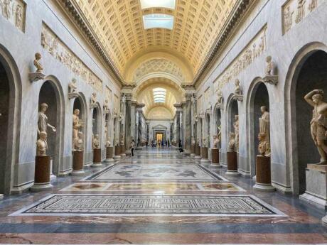 A wide marbled corridor with mosaic on the floor and statues in alcoves down the side.