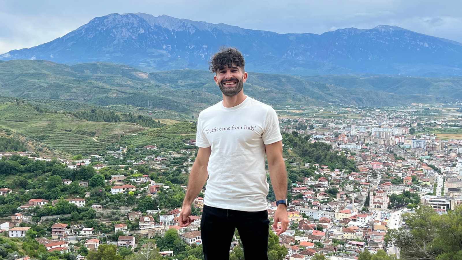 Stefan standing in front of a town and hills in the distance, wearing a white t-shirt that says "Outfit came from Italy."