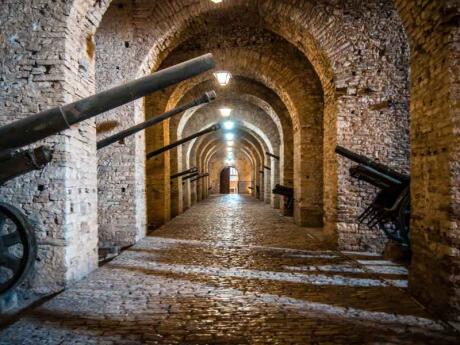 An interior castle hallway with stone arches and old cannons poking out from recesses.