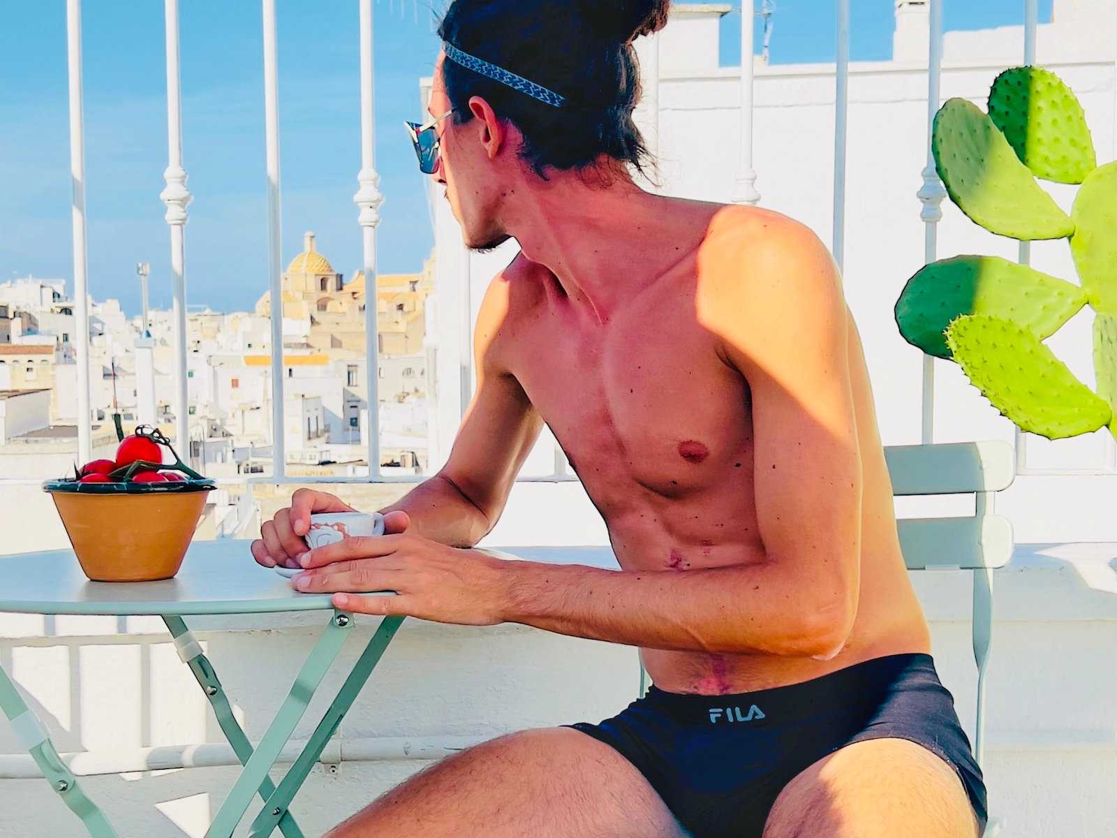 A gay man in his underpants from Italy sitting outside at a table turning away from the camera.