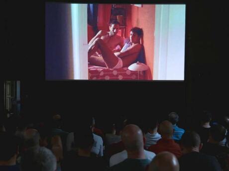The back of people's heads looking at a queer film screening with two men in bed.