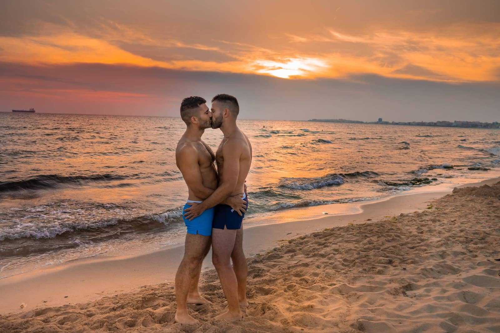 Nomadic Boys in shorts kissing on a beach in front of a sunset in italy