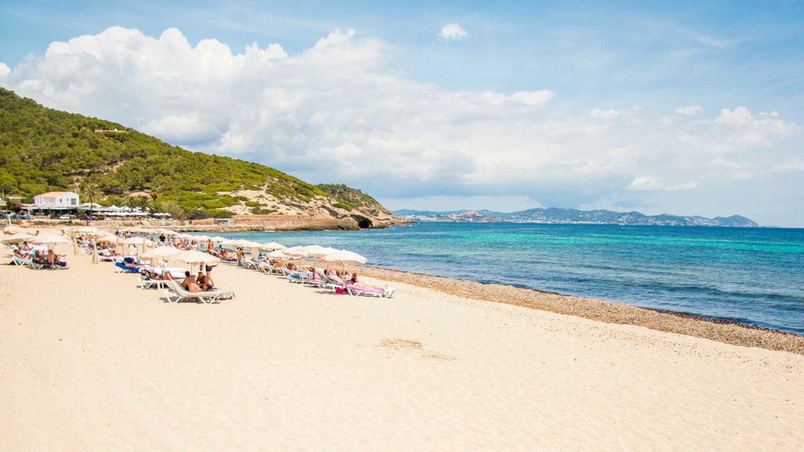 A pristine beach in Ibiza on a sunny day with men on beach lounges in the middle distance.