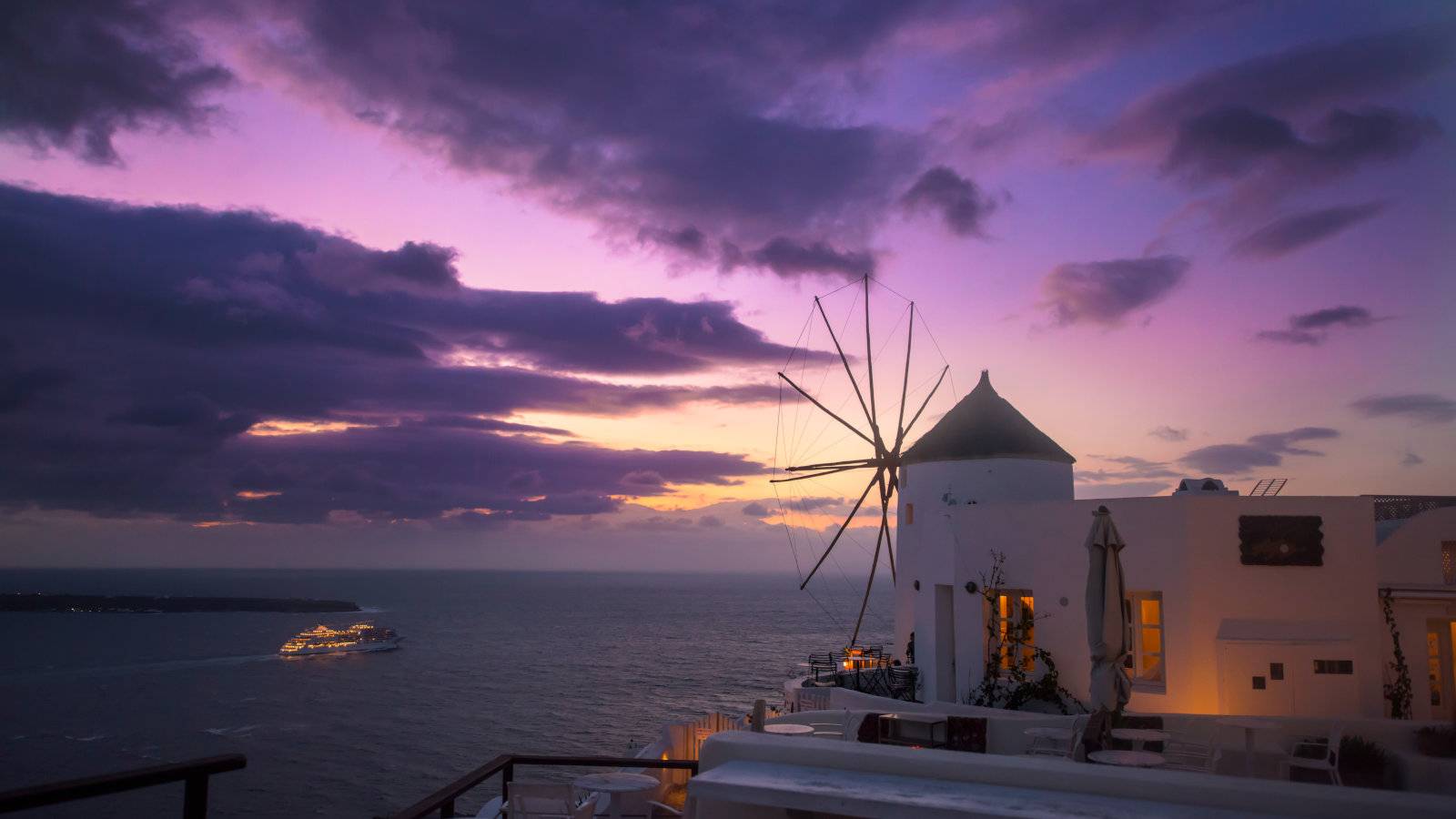 A white windmill in Santorini looking out over a cruise ship on the ocean at sunset.