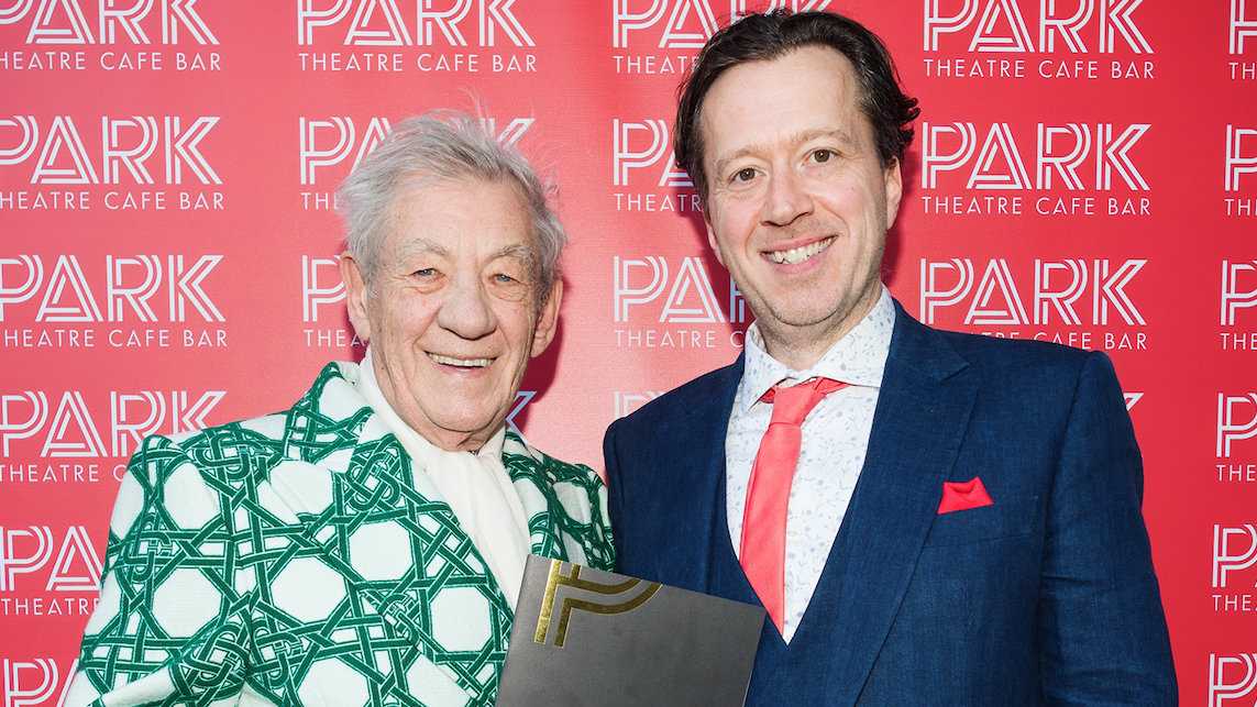 Actor Sir Ian McKellen and another man in a suit standing in front of a red background with Park Theatre Cafe Bar written on it in white.