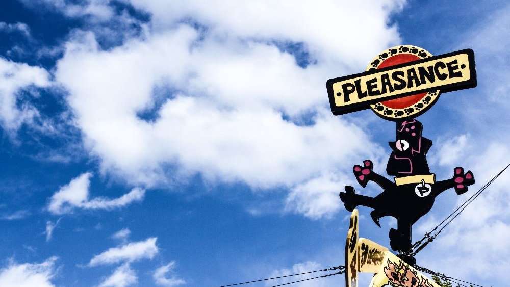 A sign with a dog balancing another sign saying "Pleasance" in front of a blue sky with fluffy white clouds.