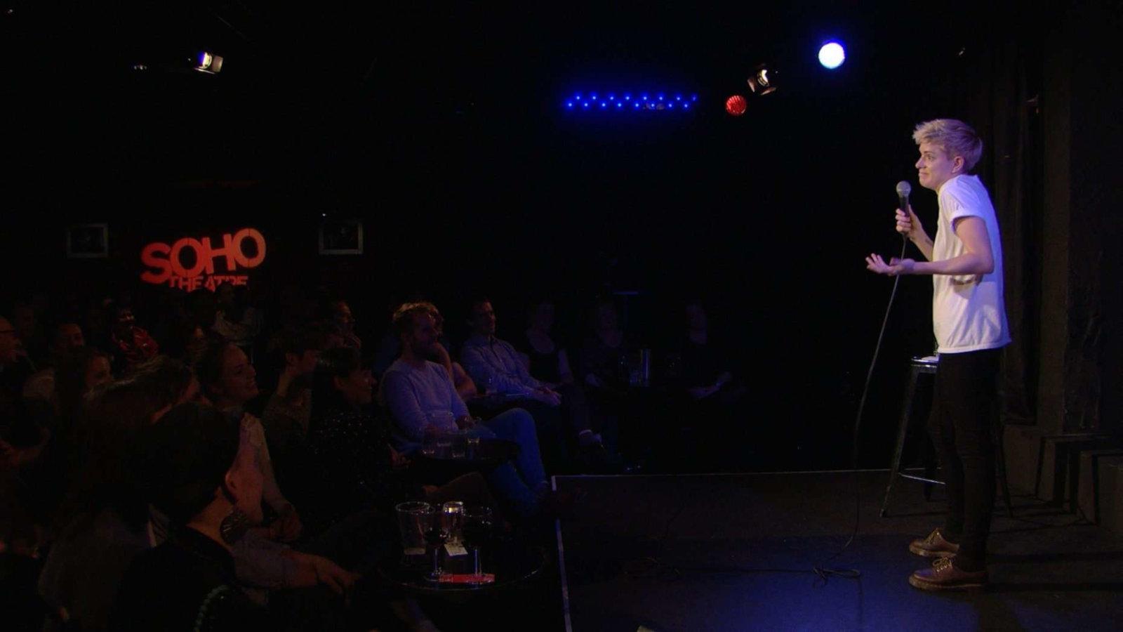 A comedian in a white t-shirt performing on stage in a dark room at the Soho Theatre in London, Soho Theatre is written on the wall in red.