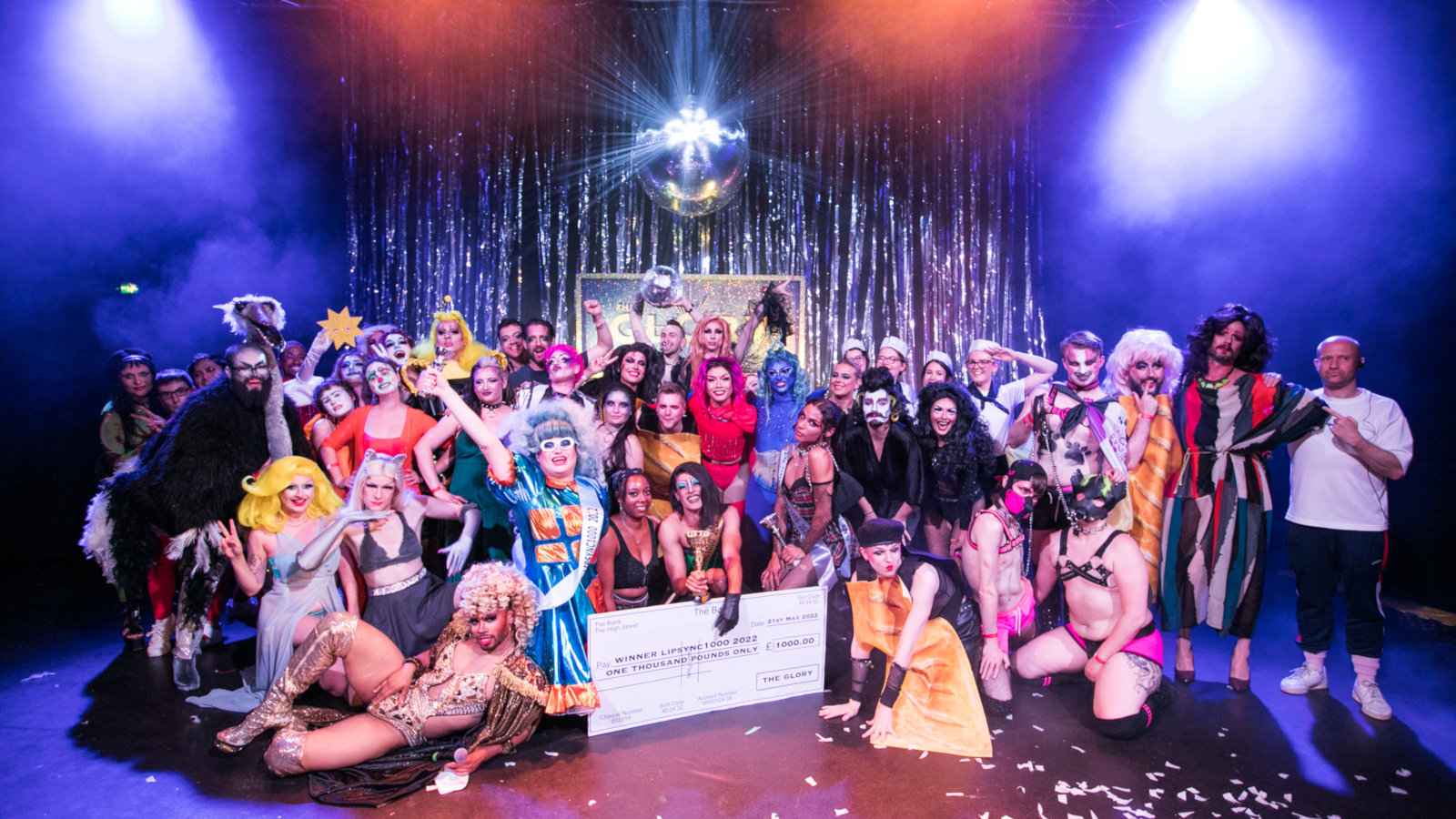 A stage with lots of people in drag and regular clothing, with lights and colored paper, as well as a giant cheque.