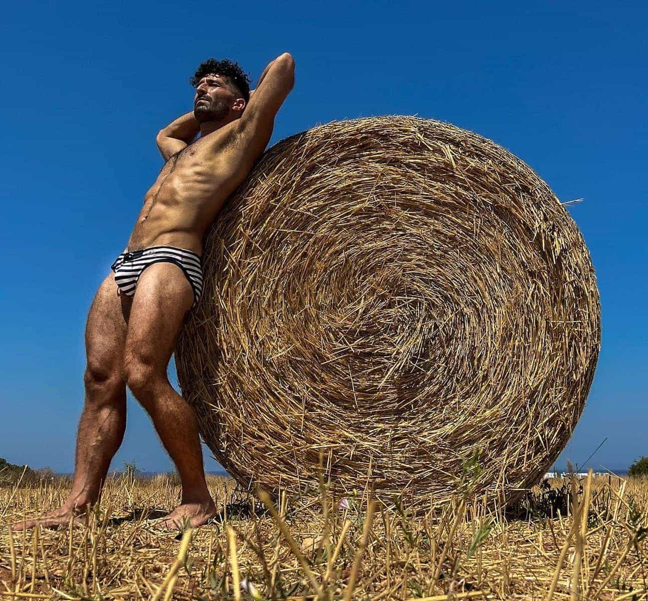 Stef in Addicted Sailor Speedos standing next to a large haystack.