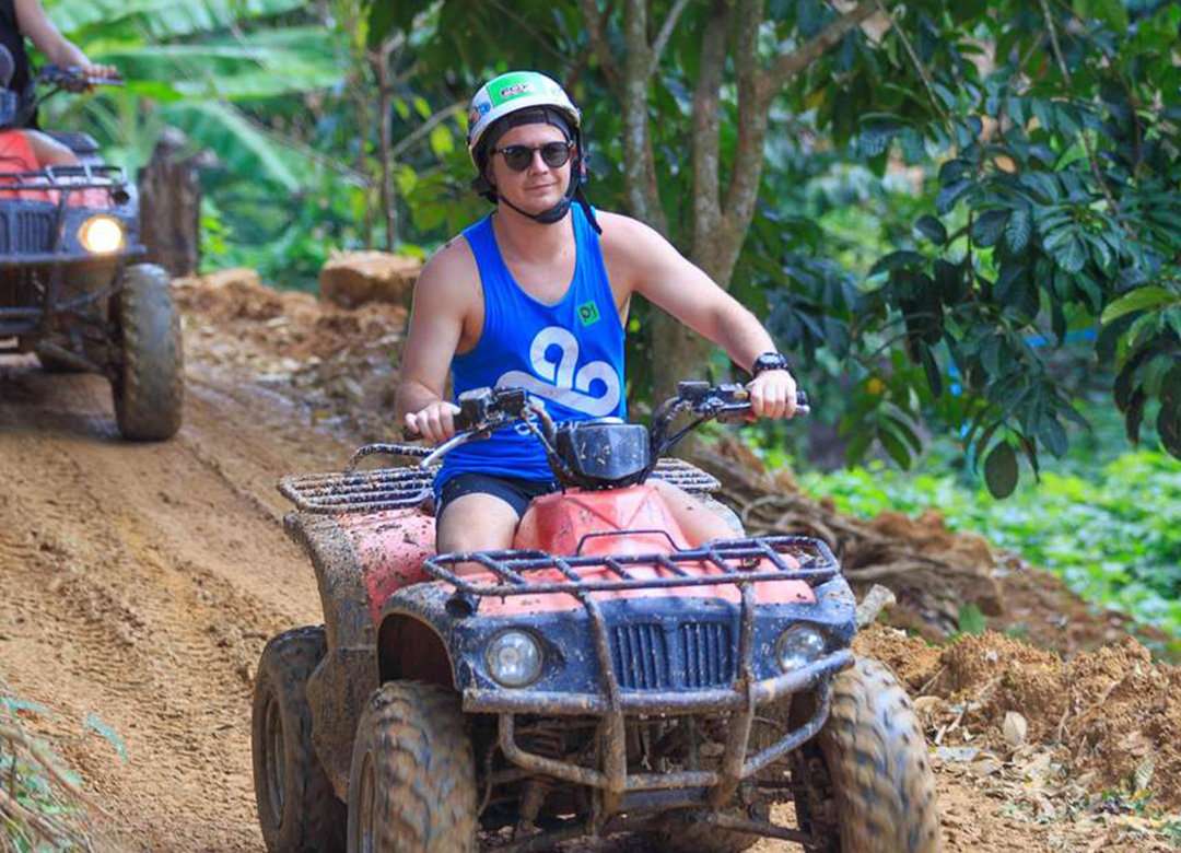 A young man in a helmet and singlet riding an ATV on a dirt path surrounded by greenery.