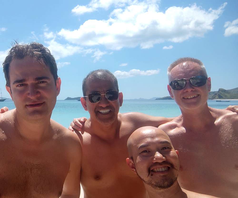 Four topless guys posing together on a beach on a sunny day and smiling.