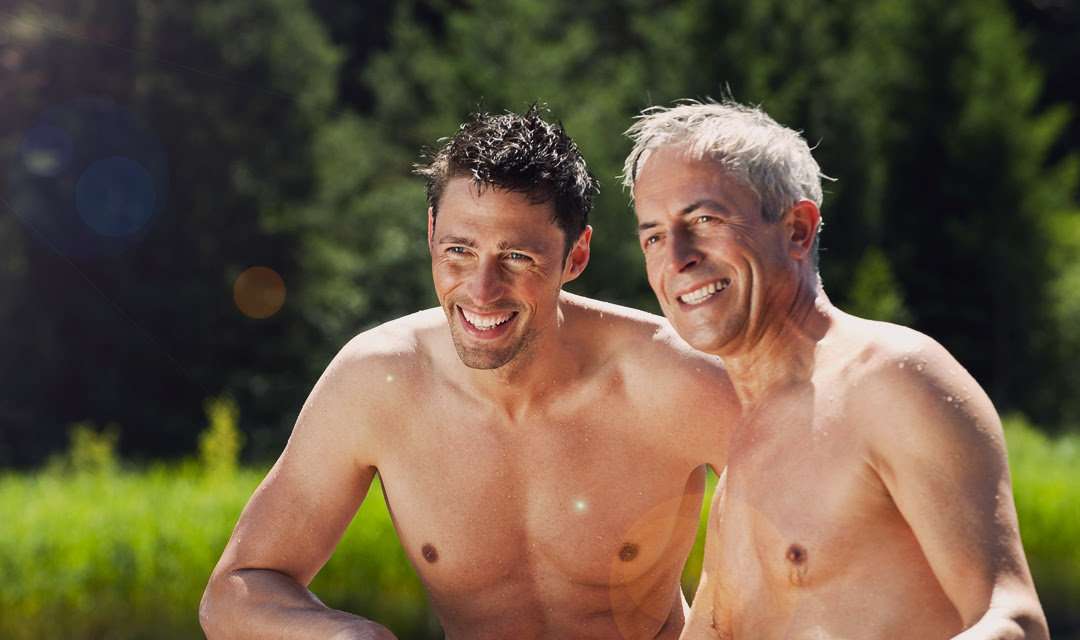 Two topless men posing together and smiling in front of some blurry greenery.