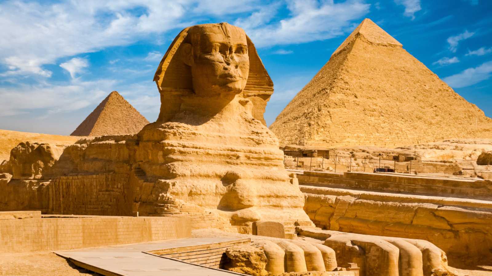 A middle distance shot of the Sphinx and pyramids in the background on a sunny day with blue sky behind the yellow structures.