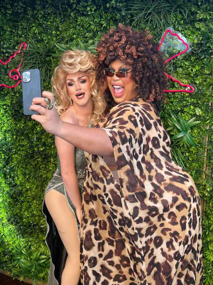 Two drag queens posing for a selfie in front of a green bush, one is blonde and not wearing much while the other has big curly hair and a big leopard print robe on.