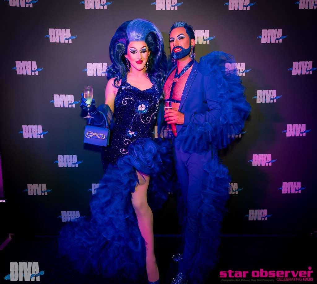 A drag queen and a man both dressed in flamboyant bright blue outfits standing in front of a backdrop with "diva" repeating on it.