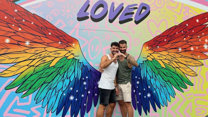 Gay couple with heart sign in front of rainbow angel wings on Oxford Street gay scene in Sydney.