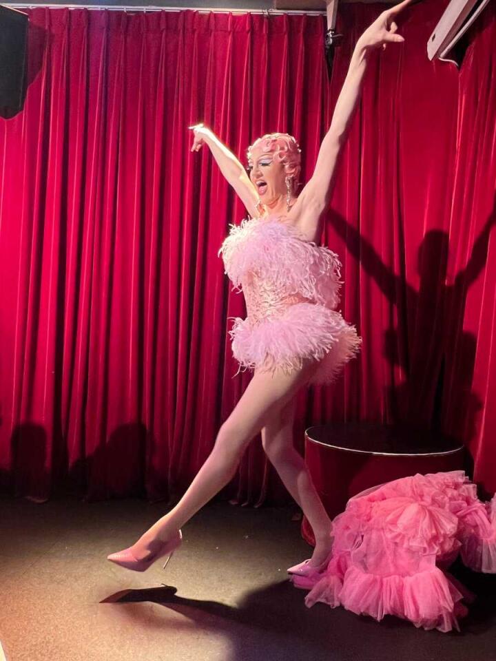 A drag queen in a cute pink dress dancing in front of a red curtain on a stage.